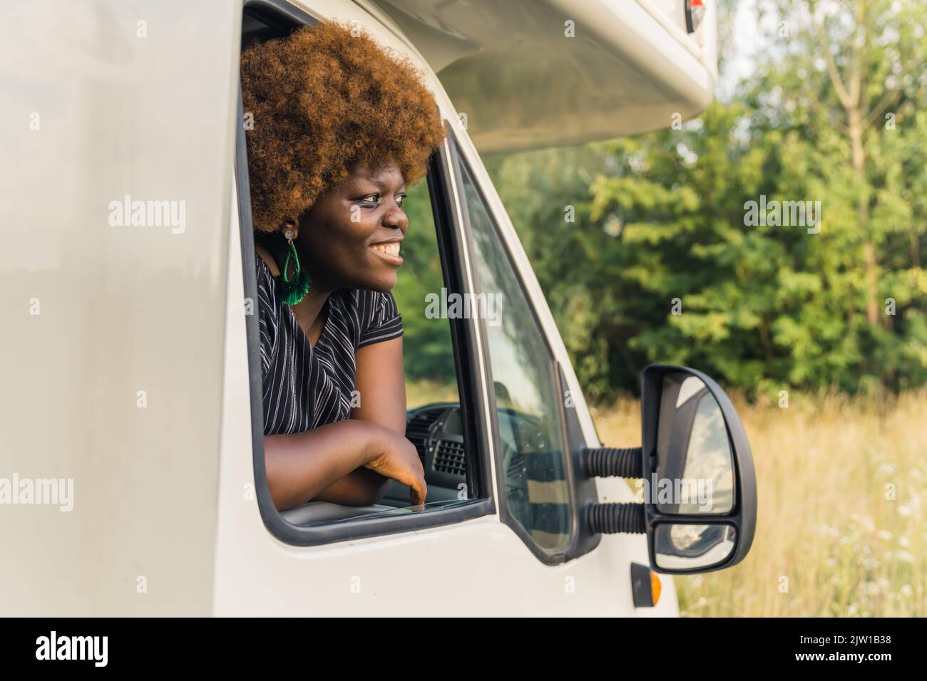 Giovane donna africana sorridente dalla pelle scura con capelli ricci che sporgono dalla finestra di un camper e che ammirano la natura pacifica intorno alla fine del suo viaggio. Foto di alta qualità Foto Stock