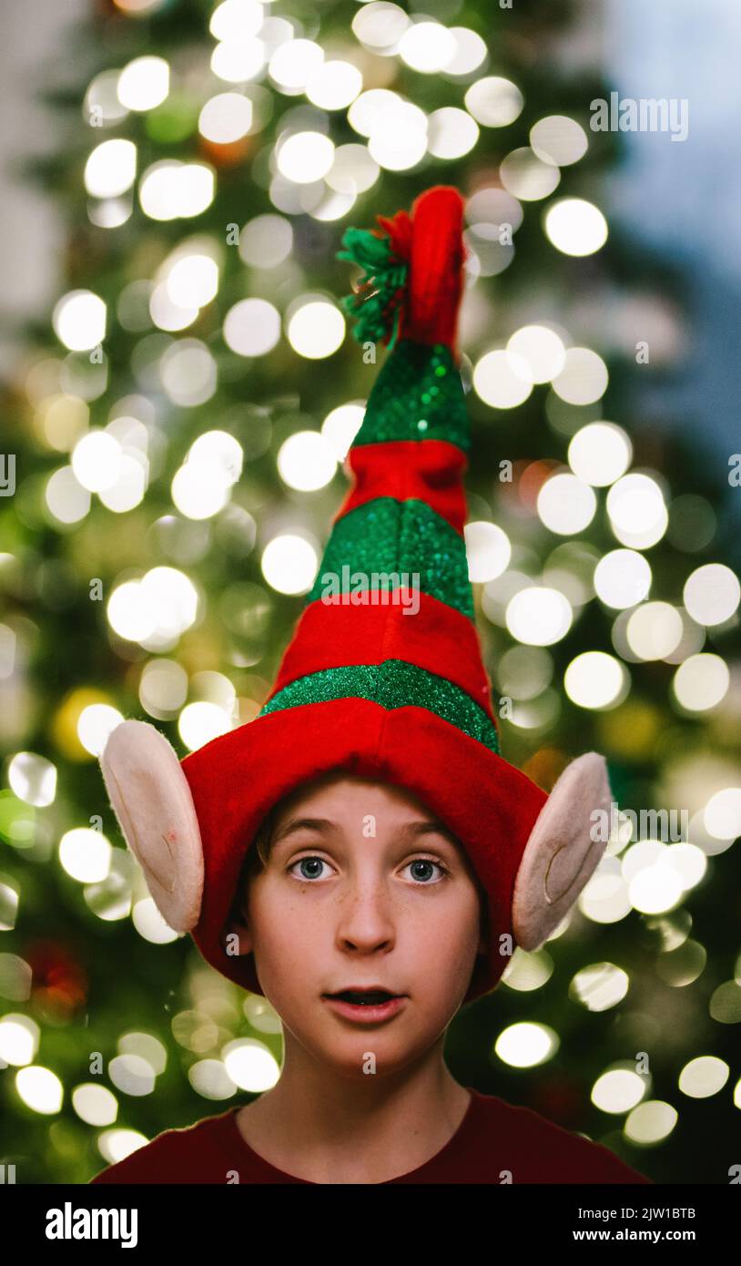 Preadolescente in cappello di elfo di natale con bokeh leggero sull'albero di natale Foto Stock