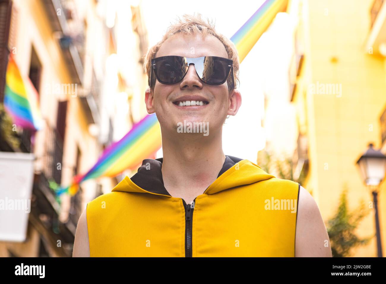 Da sotto felice non binary maschio in abiti eleganti sorridendo in occhiali da sole su sfondo sfocato di case decorate con bandiere LGBT Foto Stock