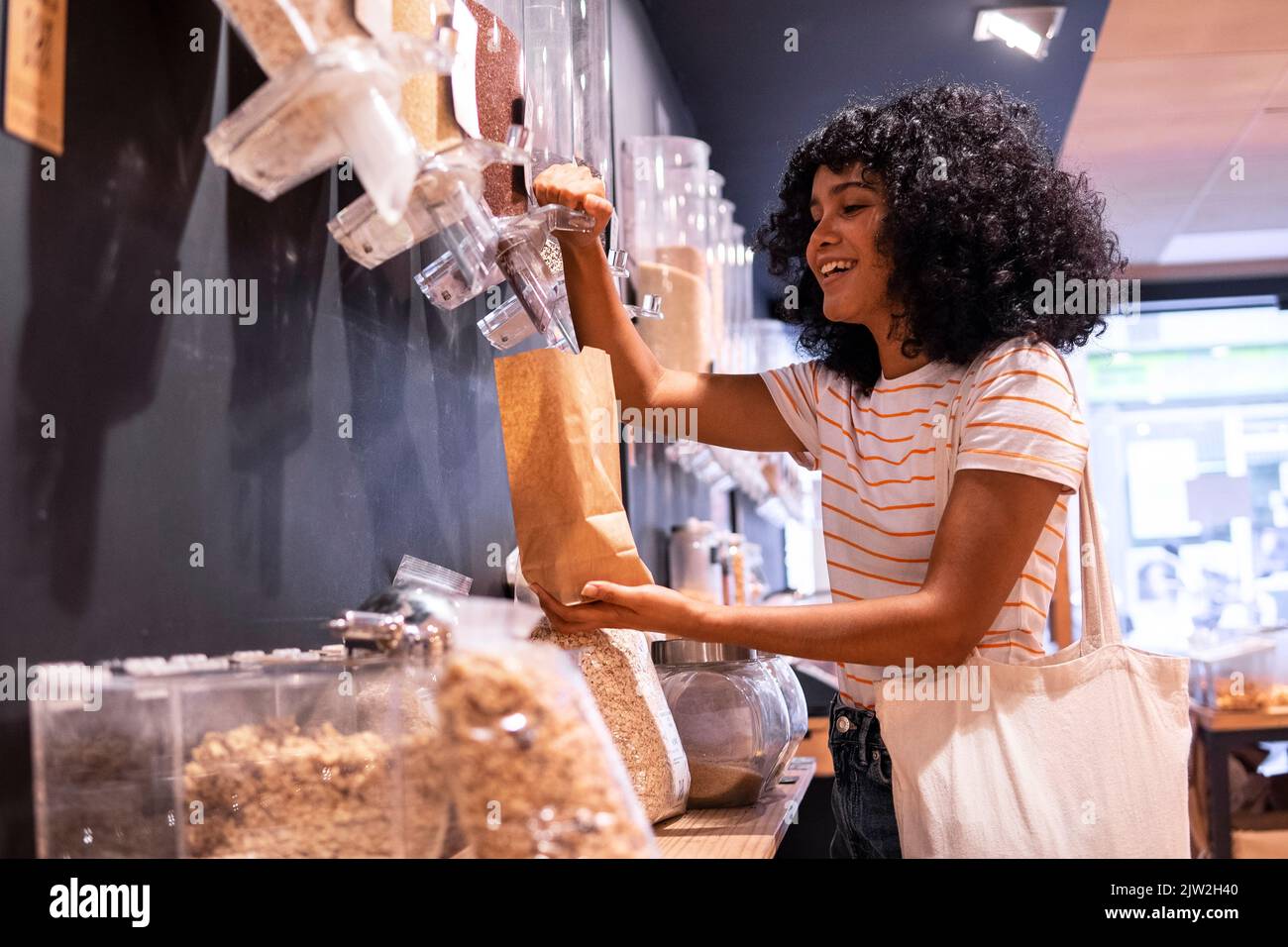 Allegra giovane afroamericana donna in abiti casual con capelli ricci sorridendo e utilizzando dispenser per riempire sacchetto di carta durante la visita eco-friendly groc Foto Stock