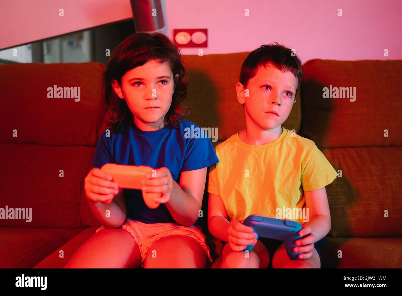 Fratelli concentrati con game pad che giocano a videogiochi mentre si siedono sul divano nel soggiorno con luce soffusa che guarda lontano Foto Stock