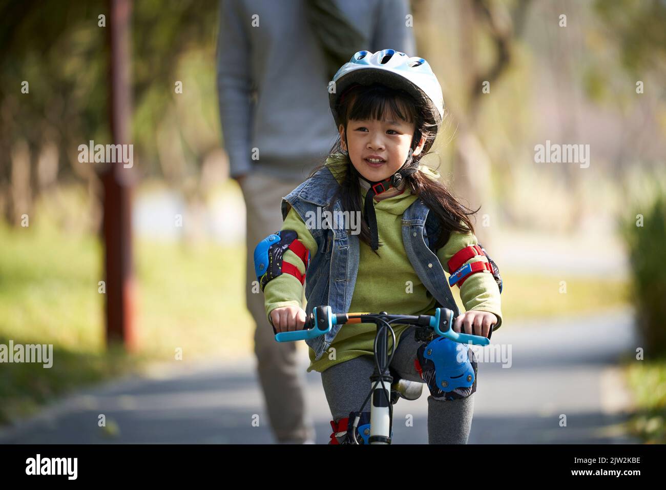bambina asiatica prescolare in bicicletta per bambini nel parco con padre che cammina dietro Foto Stock