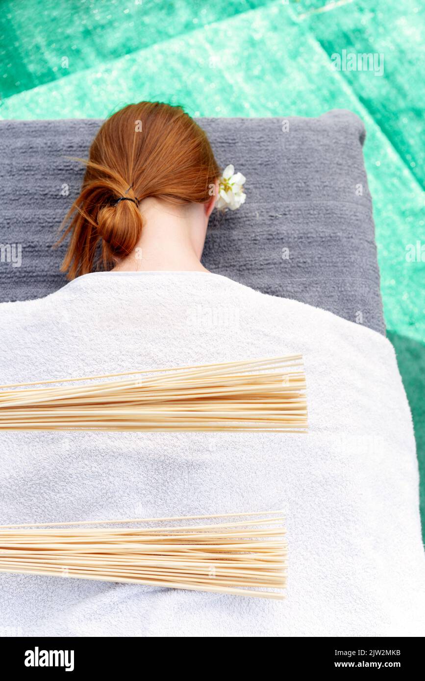 Colpisci la persona che colpisce la parte posteriore della cliente femminile rossa distesa coperta di asciugamano con le ginocine di bambù durante la sessione di massaggio nel salone di stazione termale Foto Stock