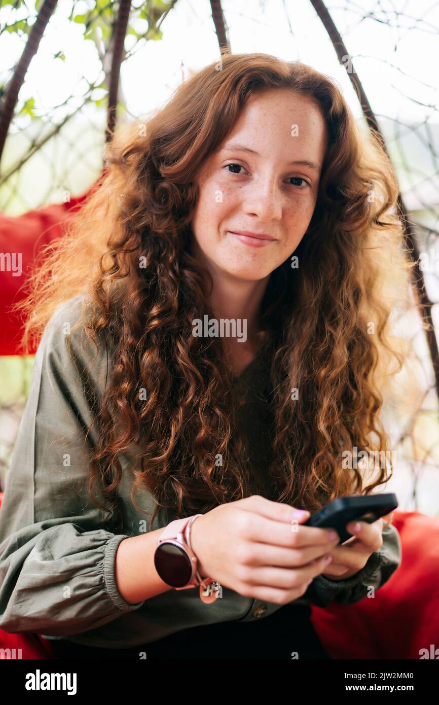 Giovane donna positiva con lunghi capelli di zenzero in abiti casual sorridendo e guardando la macchina fotografica mentre si aggrediscia su una sedia a uovo in vimini appesa utilizzando lo smartphone Foto Stock