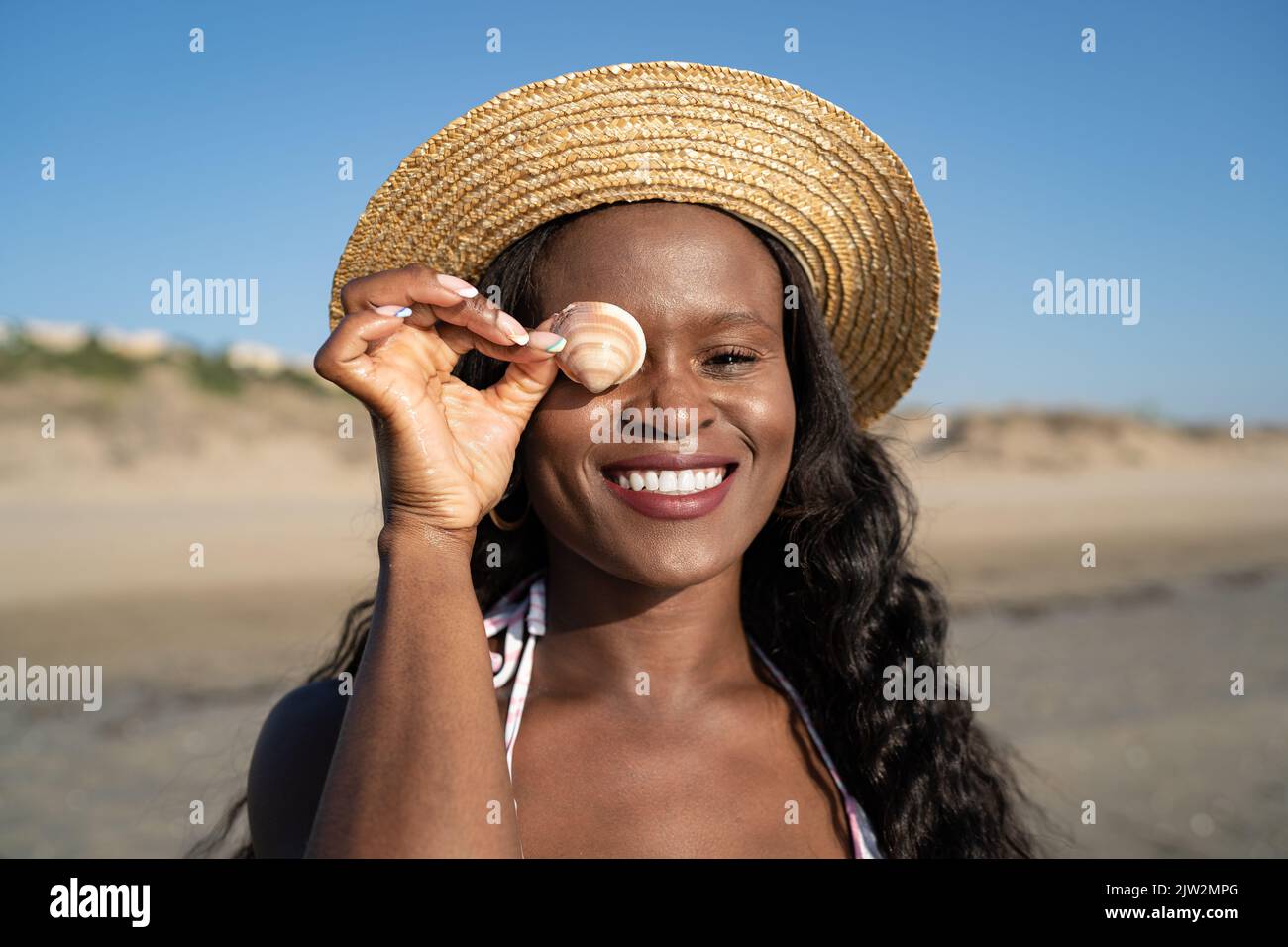 Felice ragazza afro-americana viaggiatore in cappello di paglia guardando la macchina fotografica con sorriso e occhio di copertura con conchiglia sullo sfondo sfocato della spiaggia sabbiosa A. Foto Stock