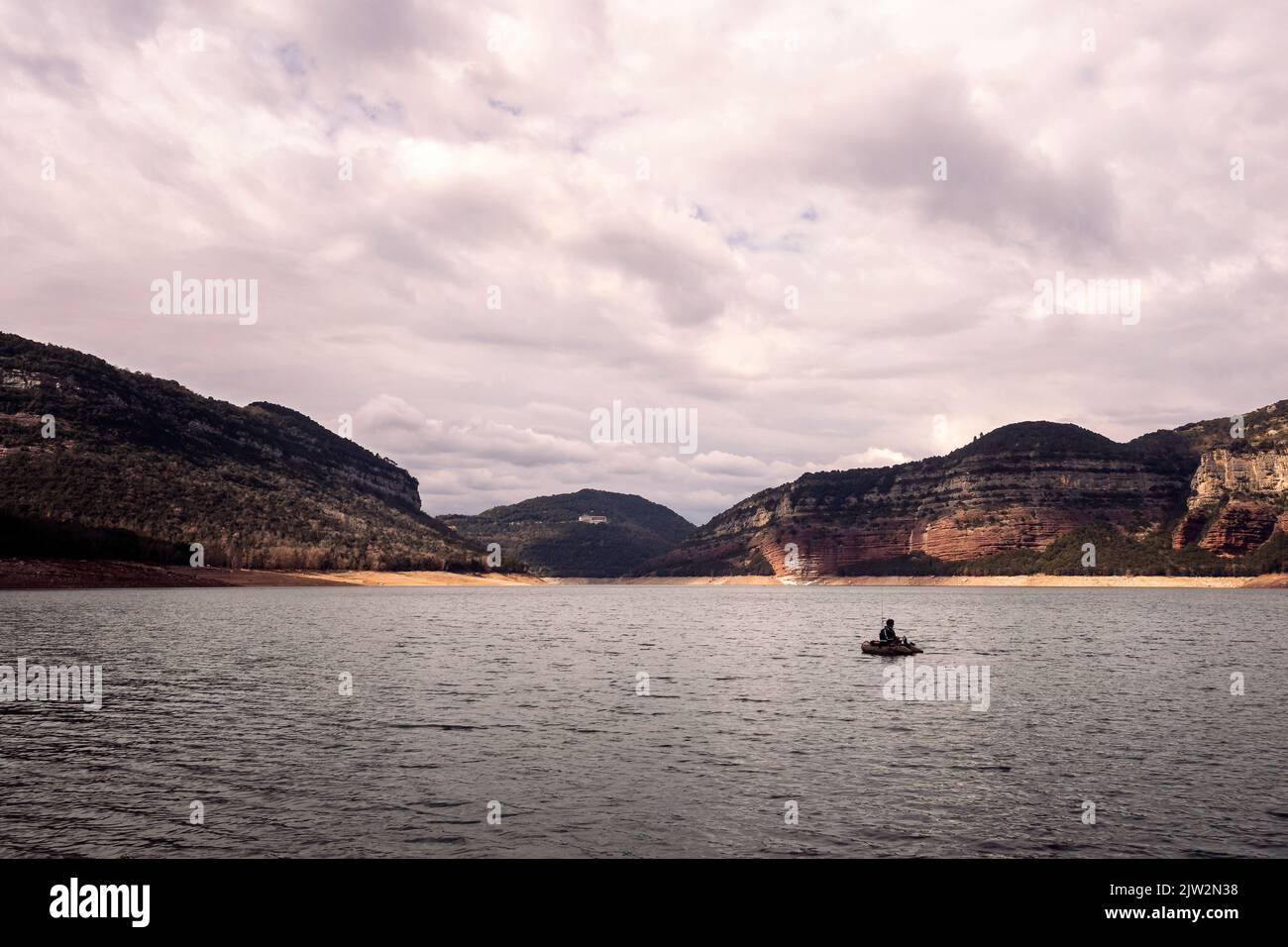 Pescatore in barca sulle acque ondulate del fiume contro le montagne e cielo nuvoloso in una giornata grigia nella natura Foto Stock