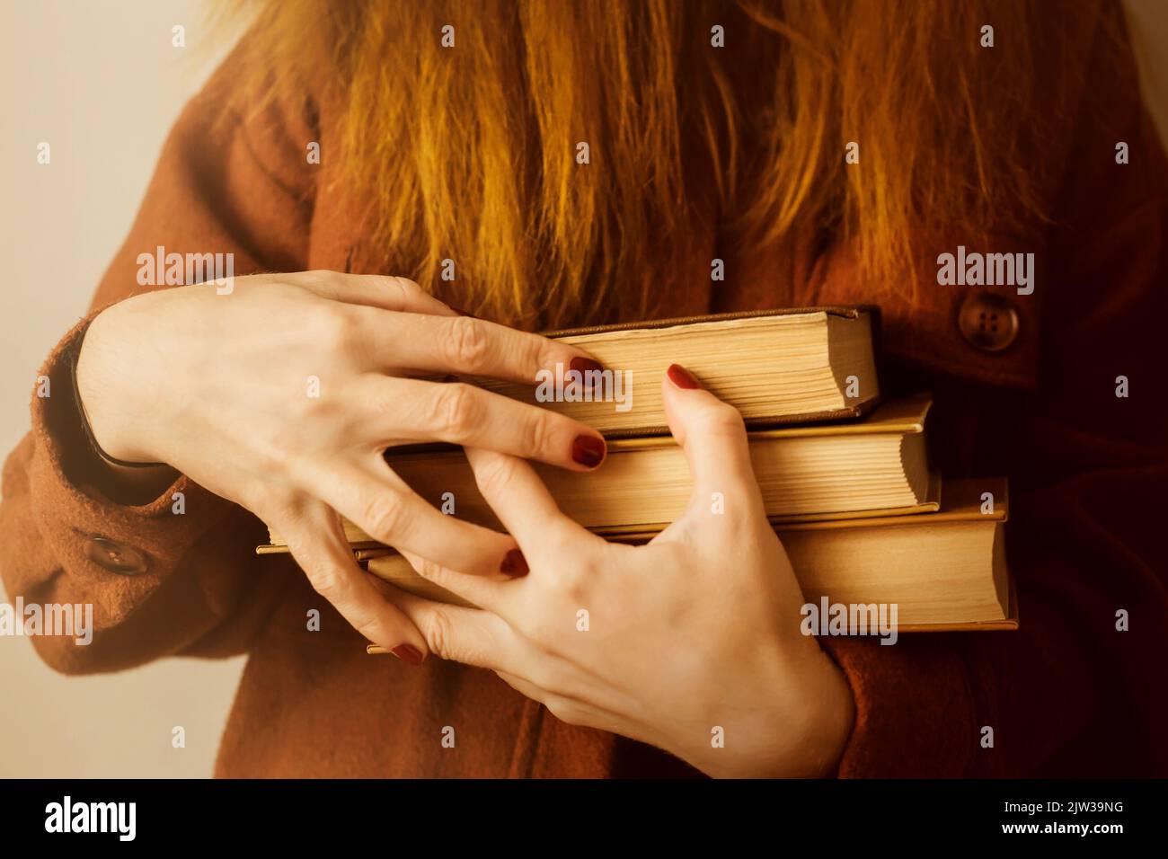 Una donna con i capelli di zenzero in una giacca di lana tiene in mano una pila di vecchi libri ricoperti di marrone. Formazione degli studenti e letteratura scientifica. Amore di r Foto Stock