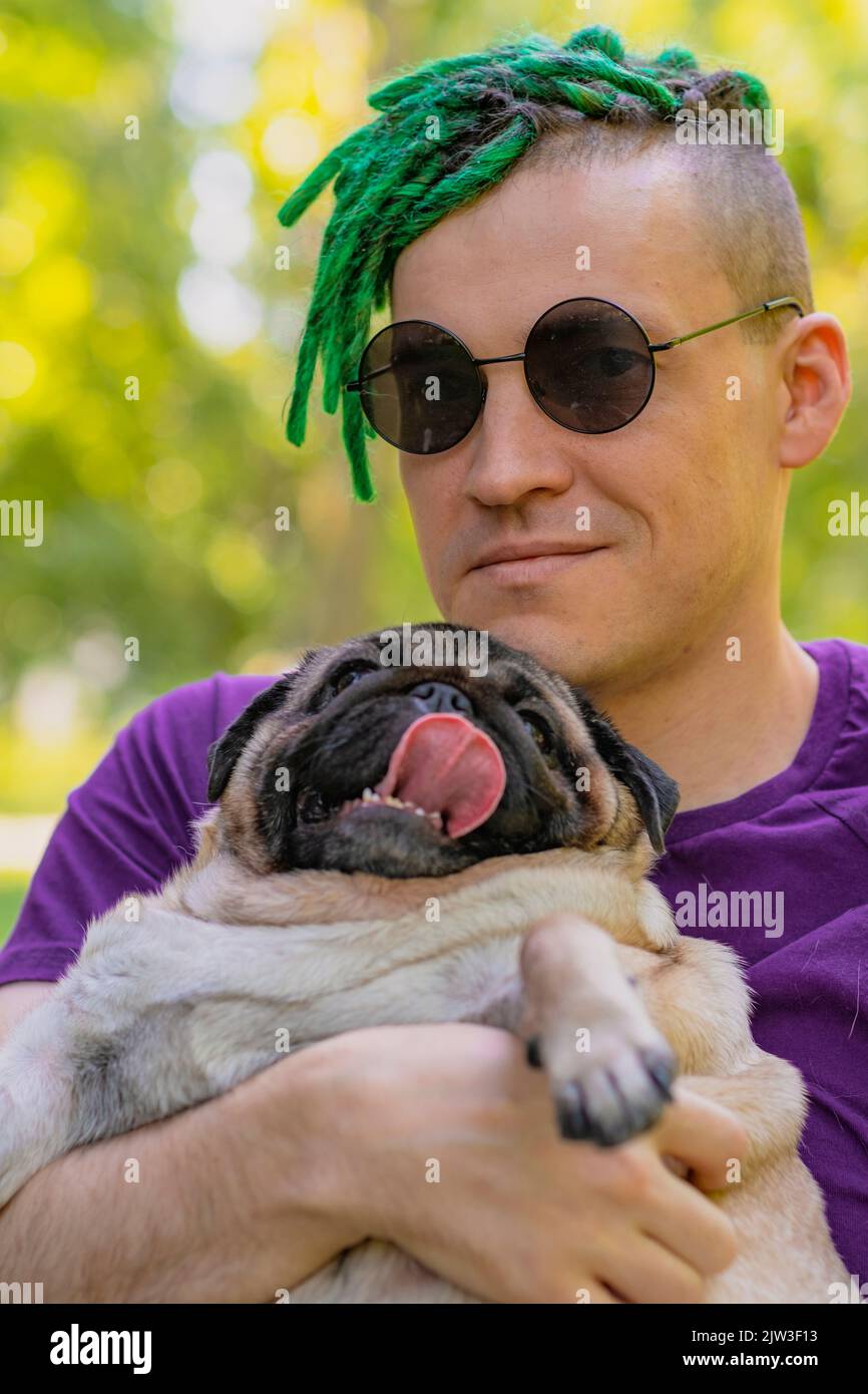 Hippster con greggi verdi e bicchieri neri rotondi, tenendo e accarezzando una carina pug in una giornata di sole estate. Un uomo cammina il suo cane. Foto Stock