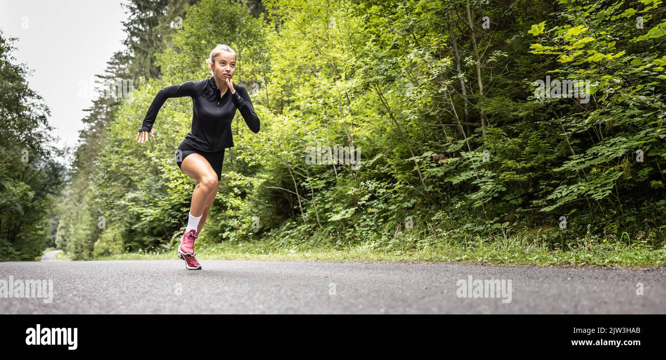 L'atleta femminile inizia con uno sprint esplosivo su una strada asfaltata nella natura. Foto Stock