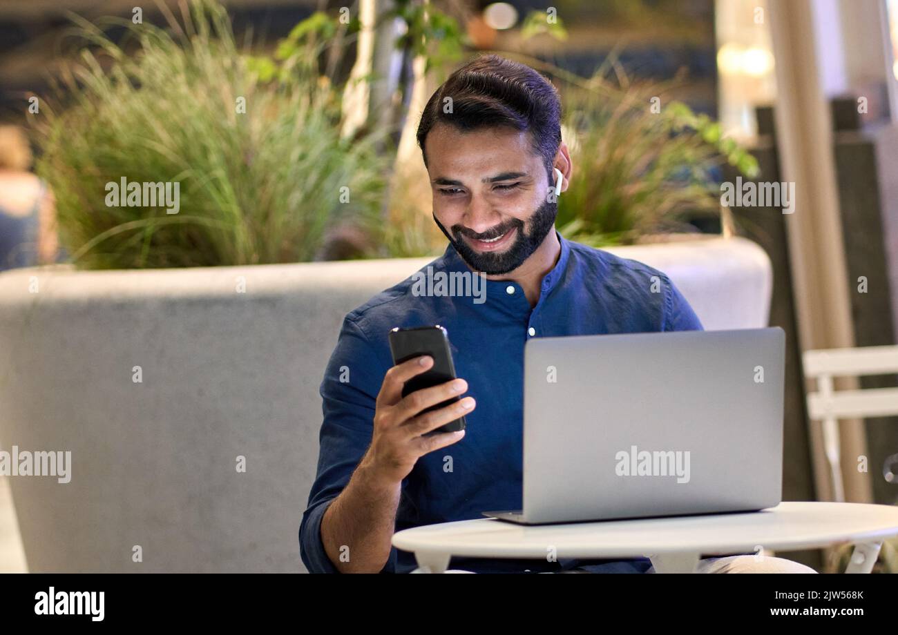 Buon uomo d'affari indiano che utilizza uno smartphone che lavora sul computer portatile di notte. Foto Stock