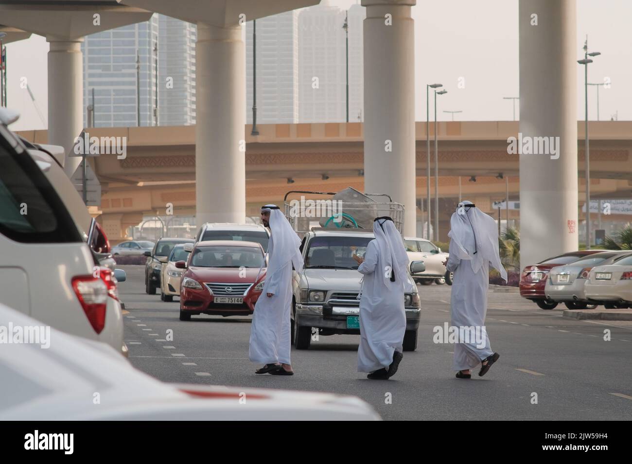 Tre uomini degli Emirati che indossano Kanduras attraversano una strada trafficata nel quartiere finanziario di Dubai, Emirati Arabi Uniti. Linea di auto che arrivano verso la gente del posto. Foto Stock
