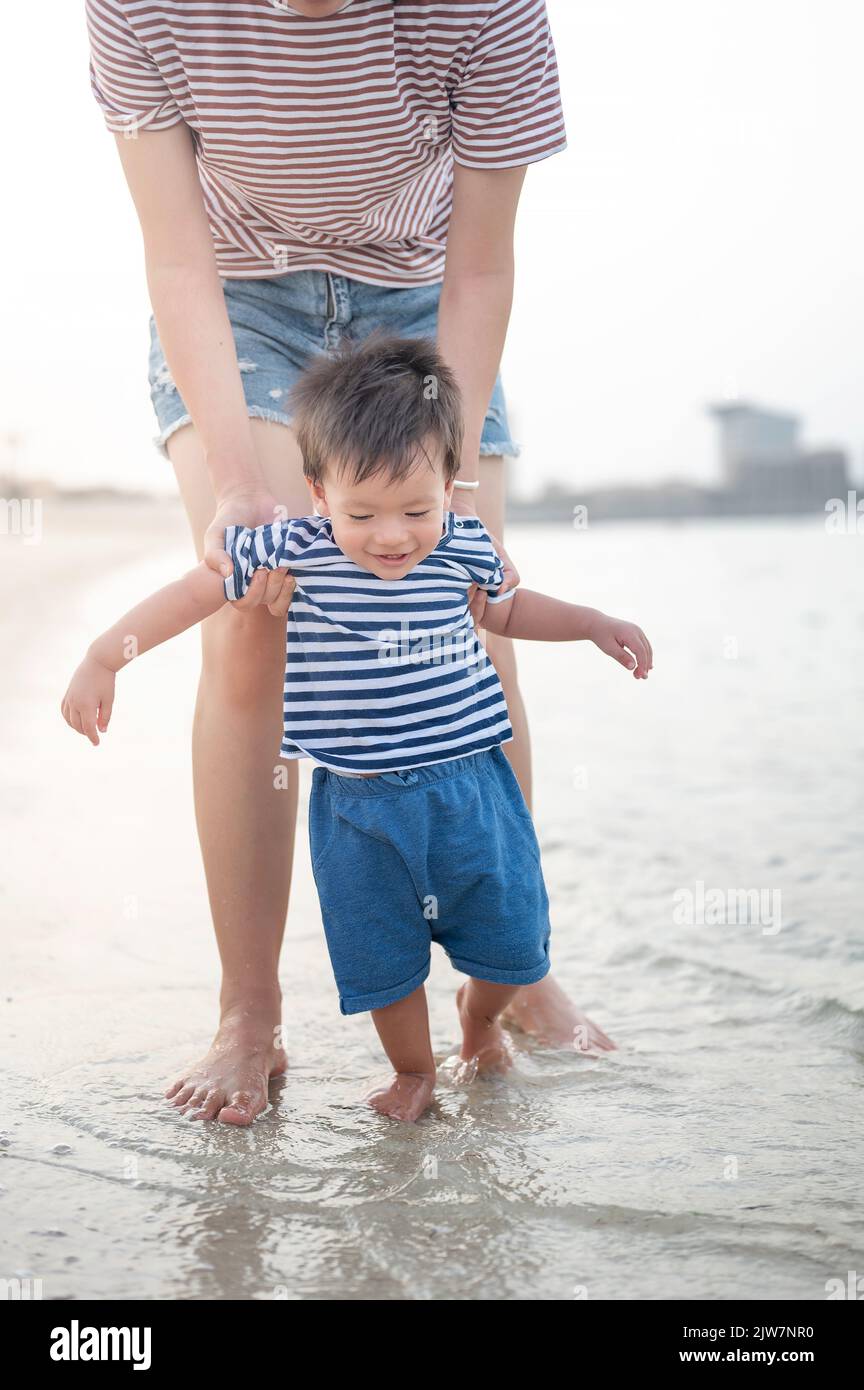 Adorabile bambino facendo i suoi primi passi sulla spiaggia in riva al mare con la madre. Un bambino di un anno imparando a camminare in una vacanza estiva Foto Stock