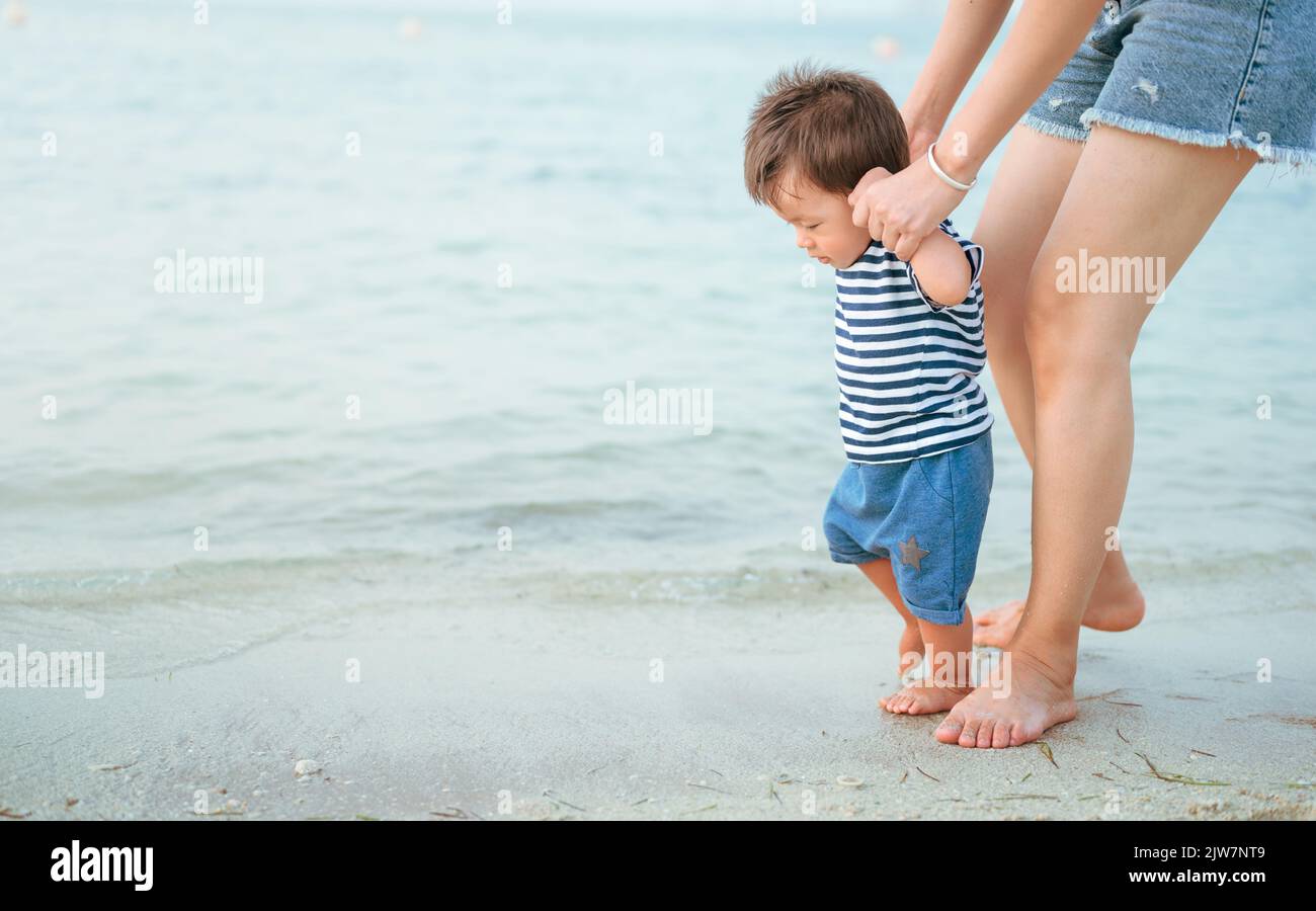Adorabile bambino facendo i suoi primi passi sulla spiaggia in riva al mare con la madre. Un bambino di un anno imparando a camminare in una vacanza estiva. L Foto Stock
