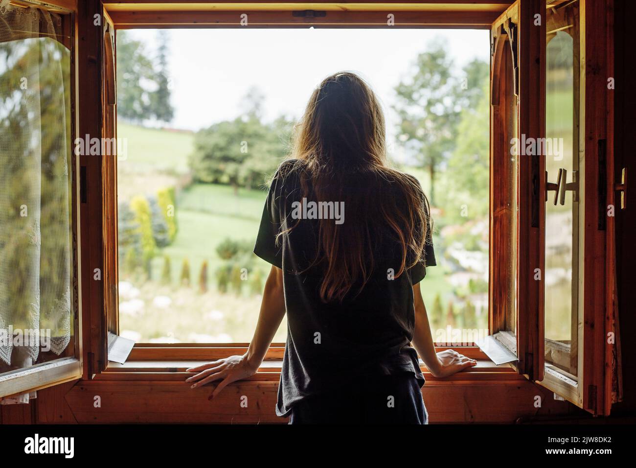 Vista posteriore di lunga donna dai capelli scuri in t shirt guardando fuori dalla finestra, contemplando paesaggio rustico albero naturale vista Foto Stock