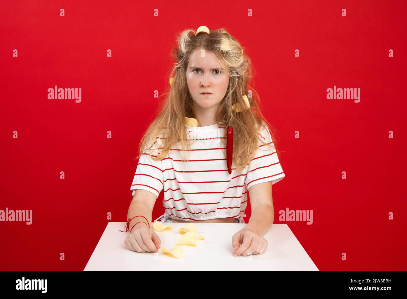 Ragazza adolescente bionda dispiaciuta, triste, offesa con bucce di agrumi arancioni e peperoncino in capelli e vestiti seduti a tavola Foto Stock