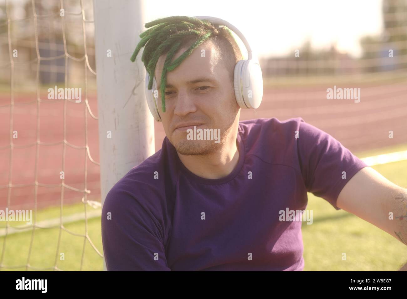 Giovane uomo con cuffie wireless che ascolta musica, chiude gli occhi dal sole luminoso, seduto sul campo di calcio, appoggiato sui cancelli Foto Stock