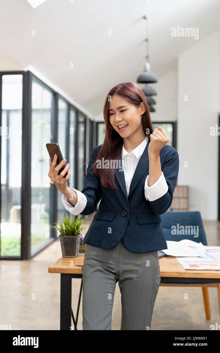 Ritratto entusiasta asiatico business donna sventolando pugni, ha ottenuto buone notizie su smartphone mentre lavorano in ufficio moderno. Foto Stock