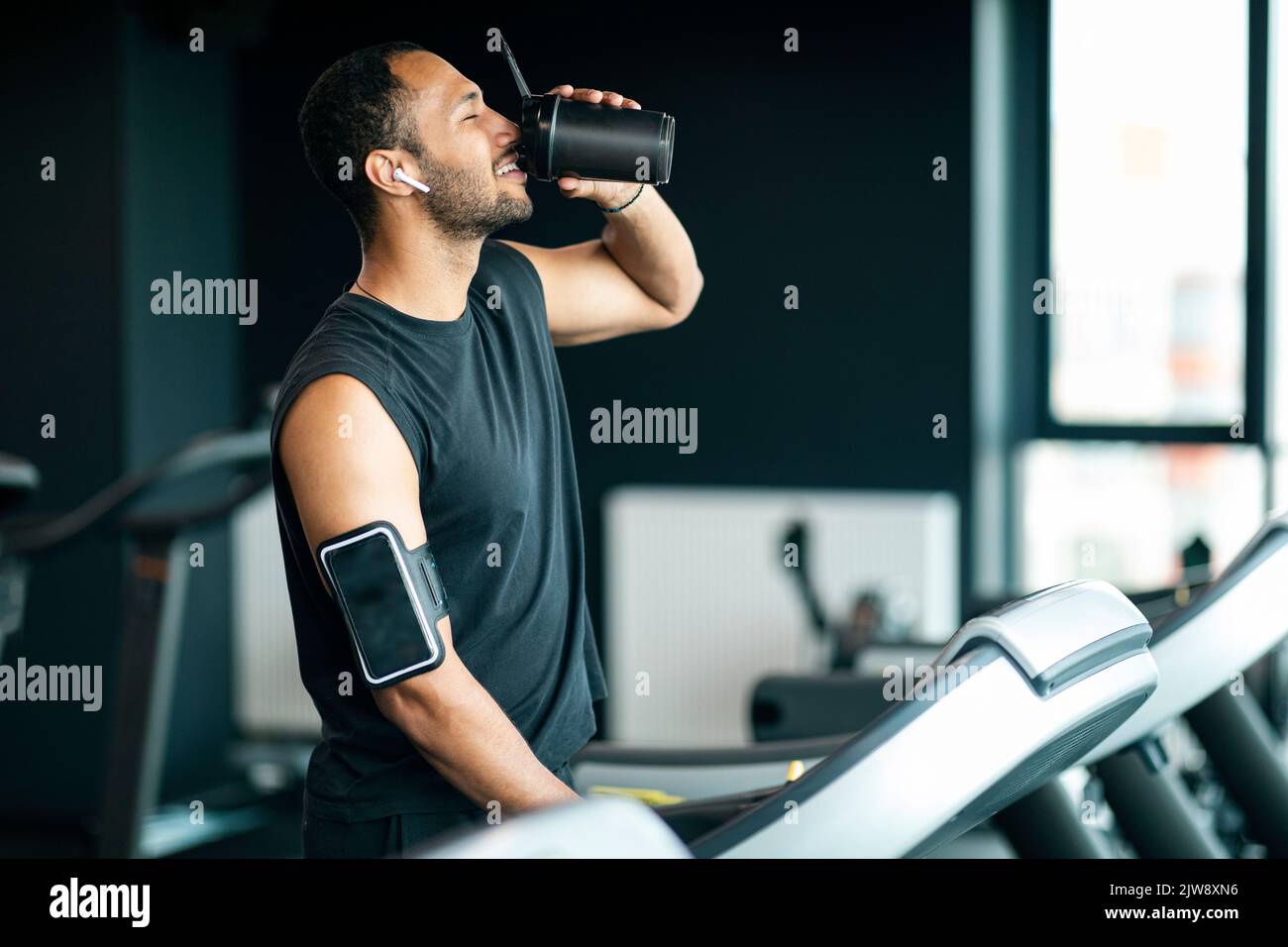 L'uomo nero beve dalla bottiglia dello shaker mentre fa jogging al tapis roulant della palestra Foto Stock