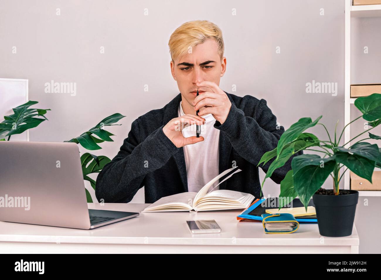 Un lavoratore dell'ufficio divertente, concentrato e arrabbiato guarda con attenzione una matita meccanica seduta alla scrivania accanto al computer portatile, ai documenti di lavoro e ai notebook. Foto Stock