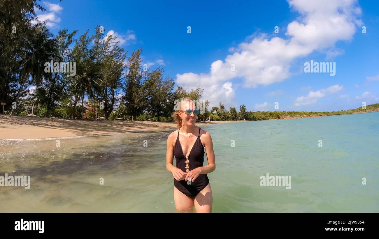 Donna bionda in costume da bagno nero in piedi in acqua sulla spiaggia tropicale Foto Stock
