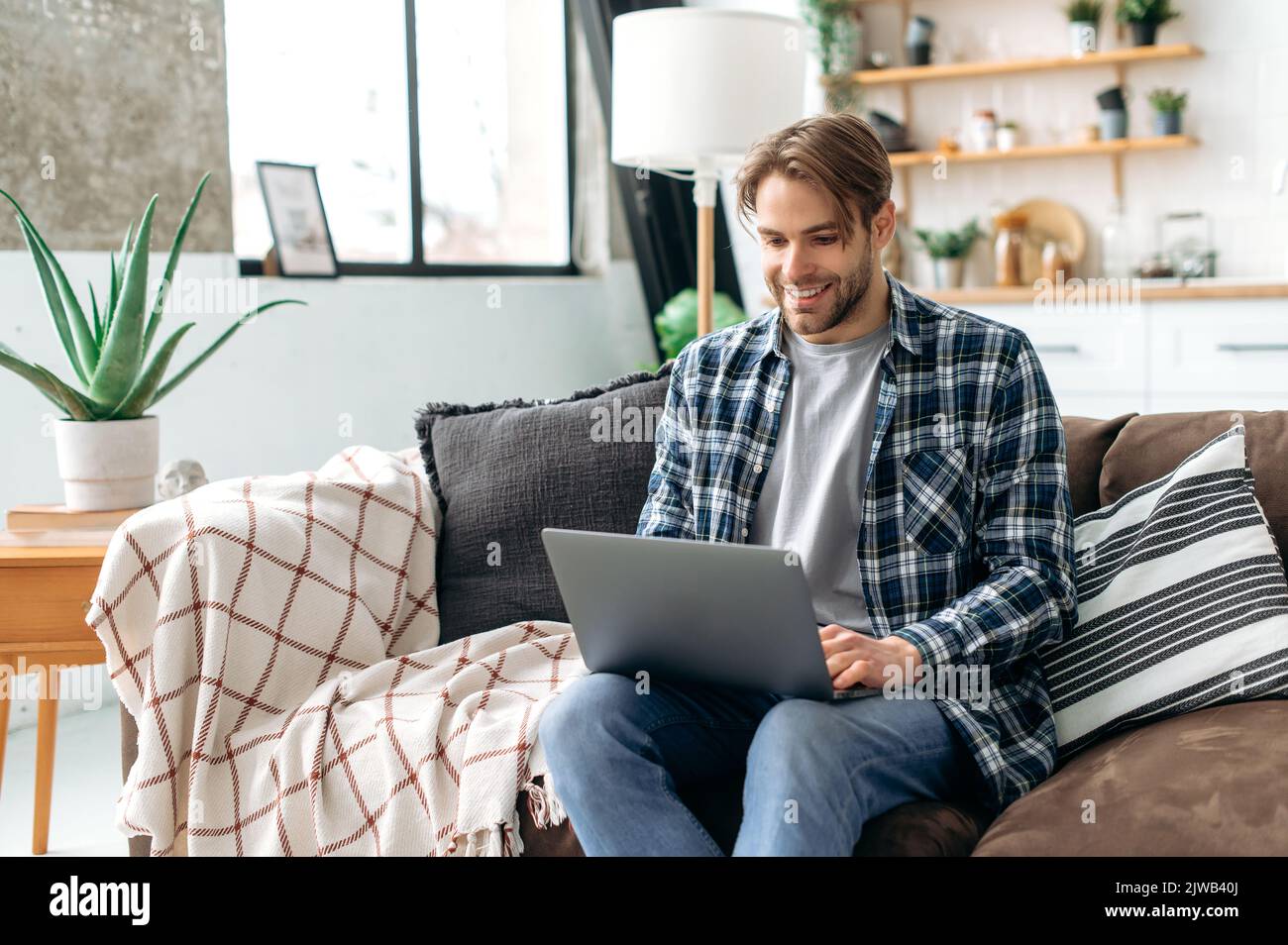 Gioioso bell'elegante caucasico di successo freelance o designer, seduto con un computer portatile su un divano nel moderno salotto mentre si lavora o studiare online, navigare informazioni in internet, sorridere Foto Stock