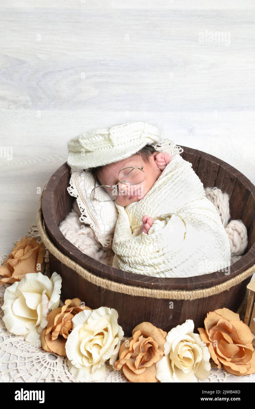 neonato carino ragazzo coperto di asciugamano e cappello dorme in legno rotondo ciotola decorativo piccoli puntelli e fiori Foto Stock