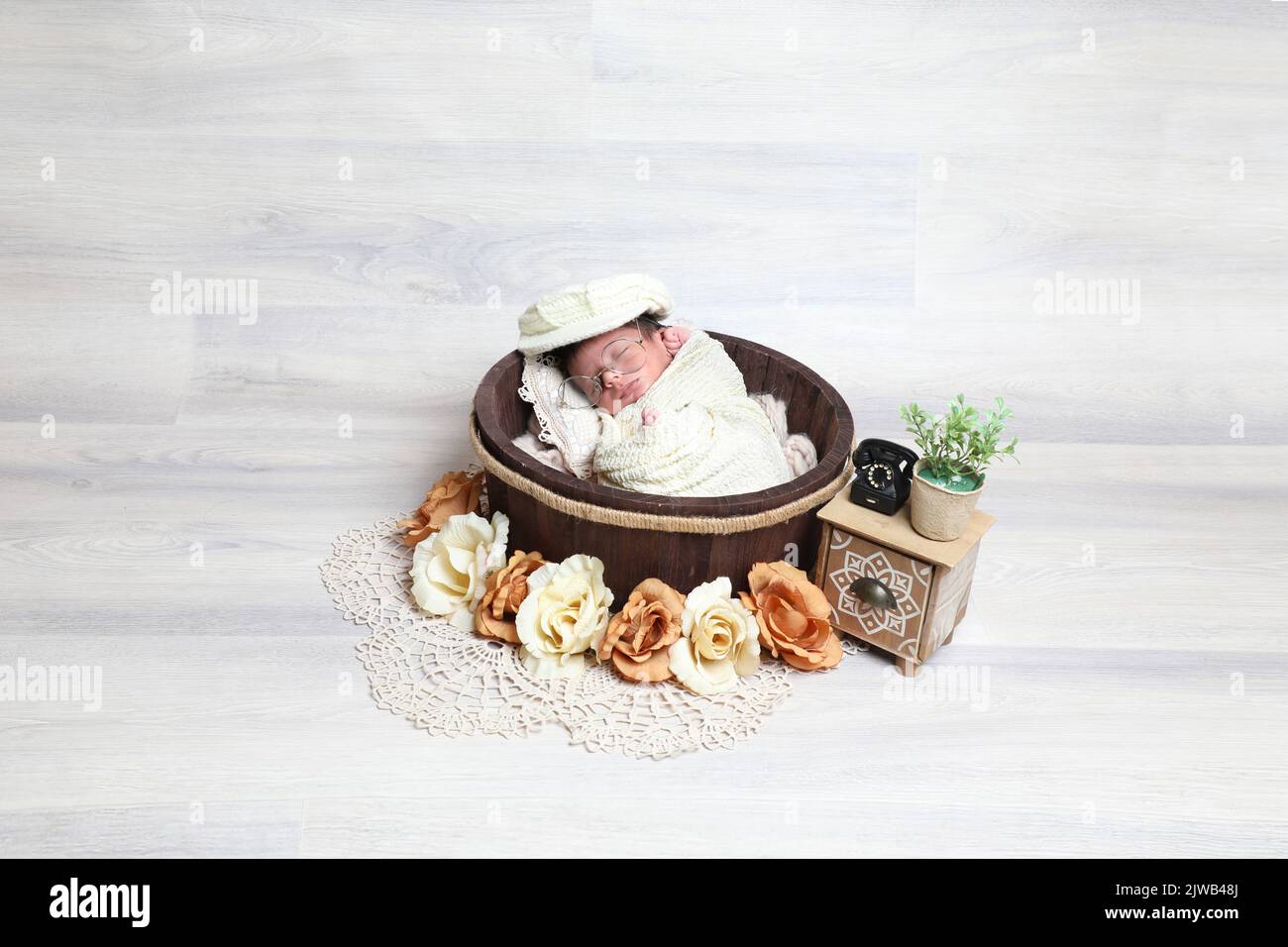 neonato carino ragazzo coperto di asciugamano e cappello dorme in legno rotondo ciotola decorativo piccoli puntelli e fiori Foto Stock