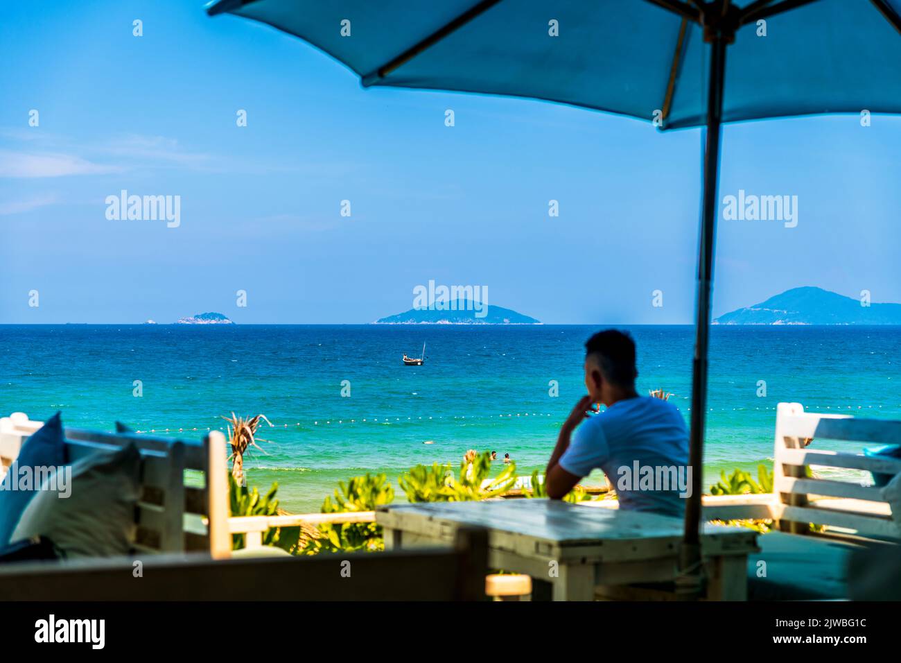Un uomo Vietnames si siede sotto un ombrellone da bar che guarda all'oceano blu di An Bang Beach, VN. Foto Stock