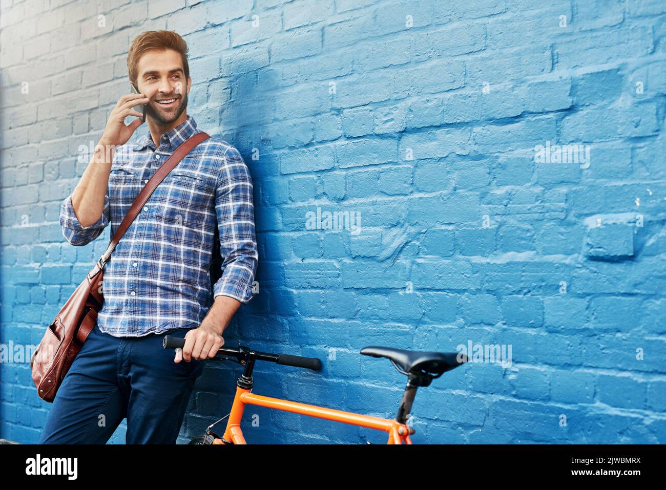 Un bel giovane che si appoggia a un muro con la sua bici che parla su un cellulare. Foto Stock