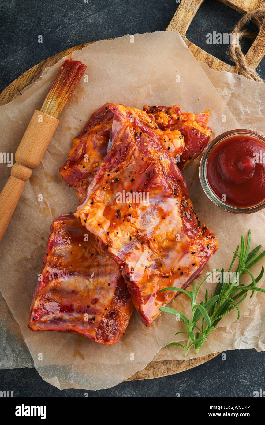 Costolette di maiale. Carne cruda. Costolette di maiale crudo in marinata con spezie, rosmarino, salsa di pomodoro e aglio su vecchio stand di legno su fondo tavolo in pietra nera. Bar Foto Stock