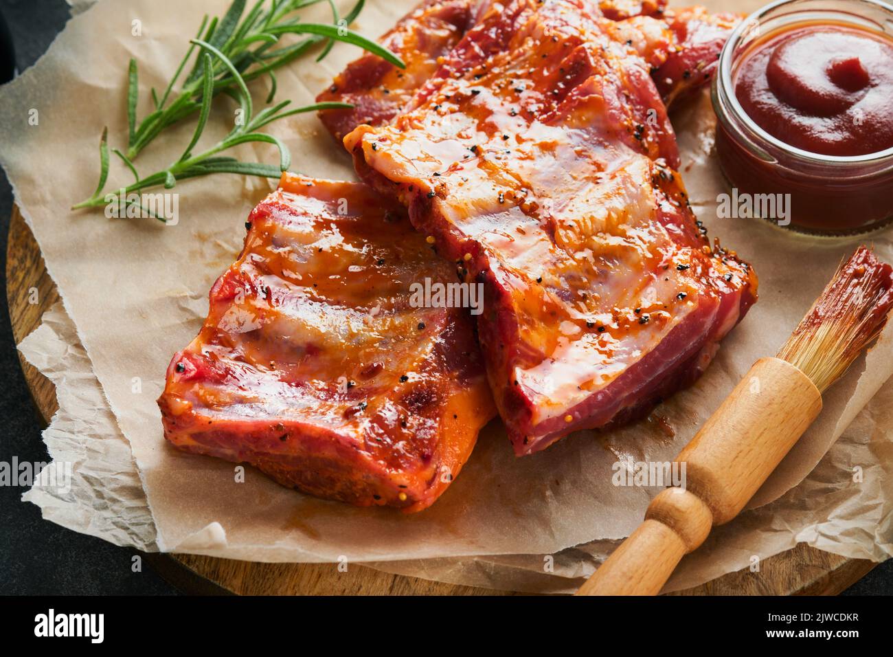 Costolette di maiale. Carne cruda. Costolette di maiale crudo in marinata con spezie, rosmarino, salsa di pomodoro e aglio su vecchio stand di legno su fondo tavolo in pietra nera. Bar Foto Stock