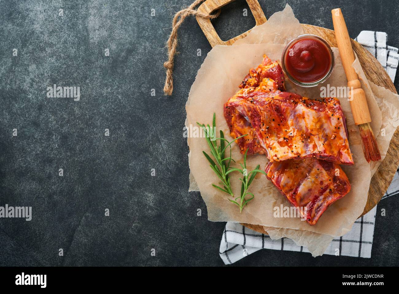Costolette di maiale. Carne cruda. Costolette di maiale crudo in marinata con spezie, rosmarino, salsa di pomodoro e aglio su vecchio stand di legno su fondo tavolo in pietra nera. Bar Foto Stock