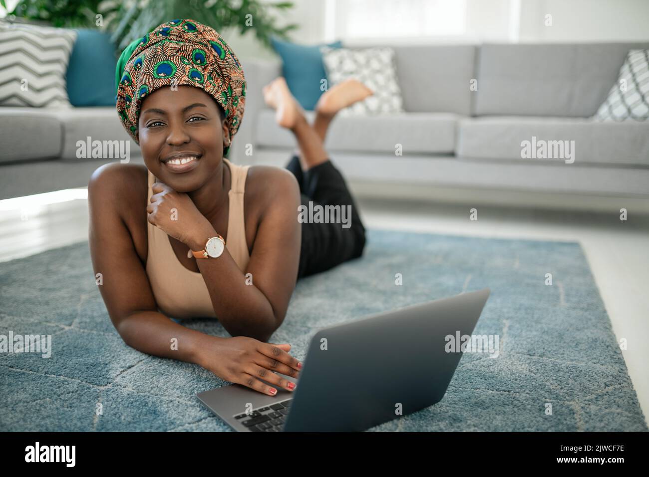Sorridente giovane ragazza africana am seduta con le gambe incrociate sul tappeto a casa utilizzando il computer portatile e guardando in macchina fotografica Foto Stock