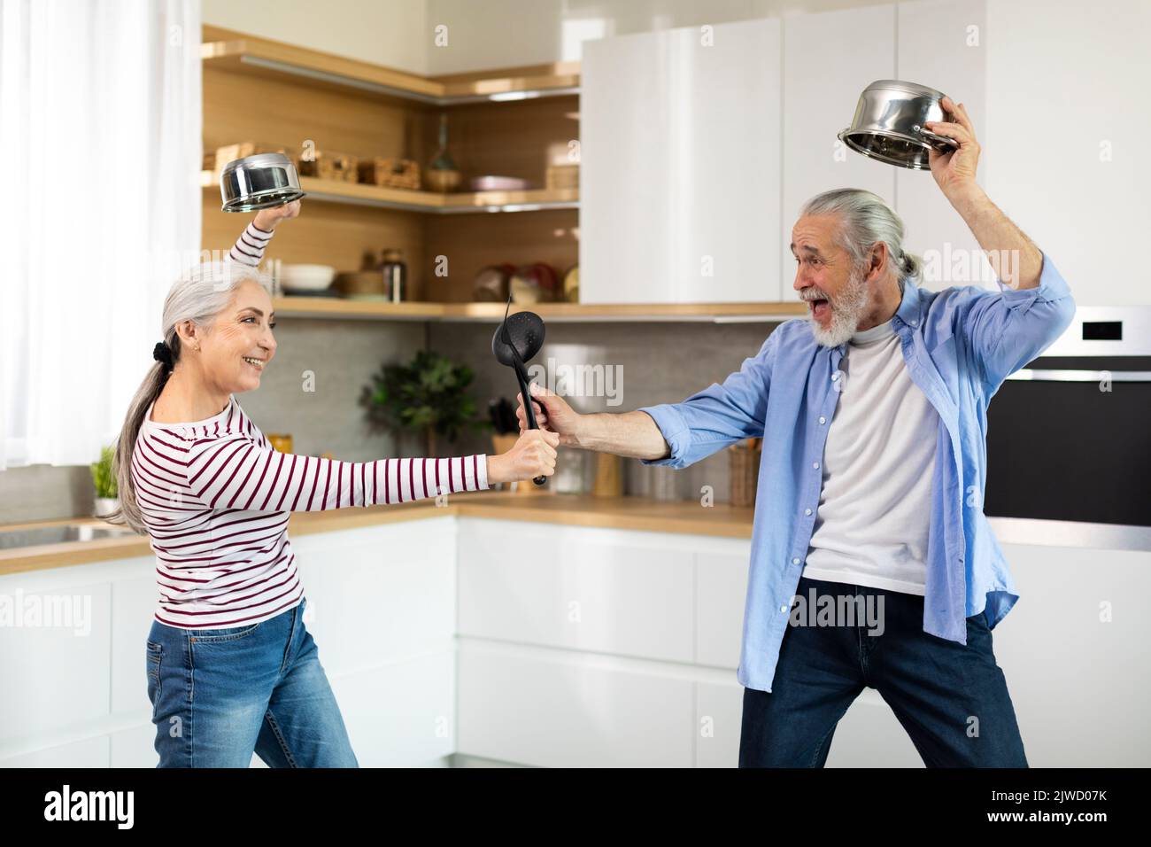 Gioioso sposi anziani divertirsi insieme in cucina, giocosamente combattendo con utensili da cucina Foto Stock