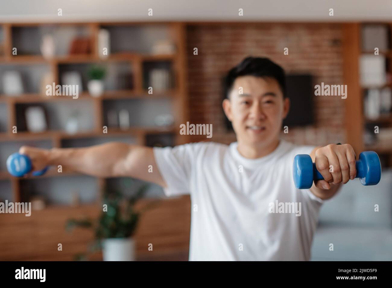 Uomo sportivo coreano maturo che si esercita con manubri a casa, rinforzando i muscoli del braccio, fuoco selettivo sulla mano Foto Stock