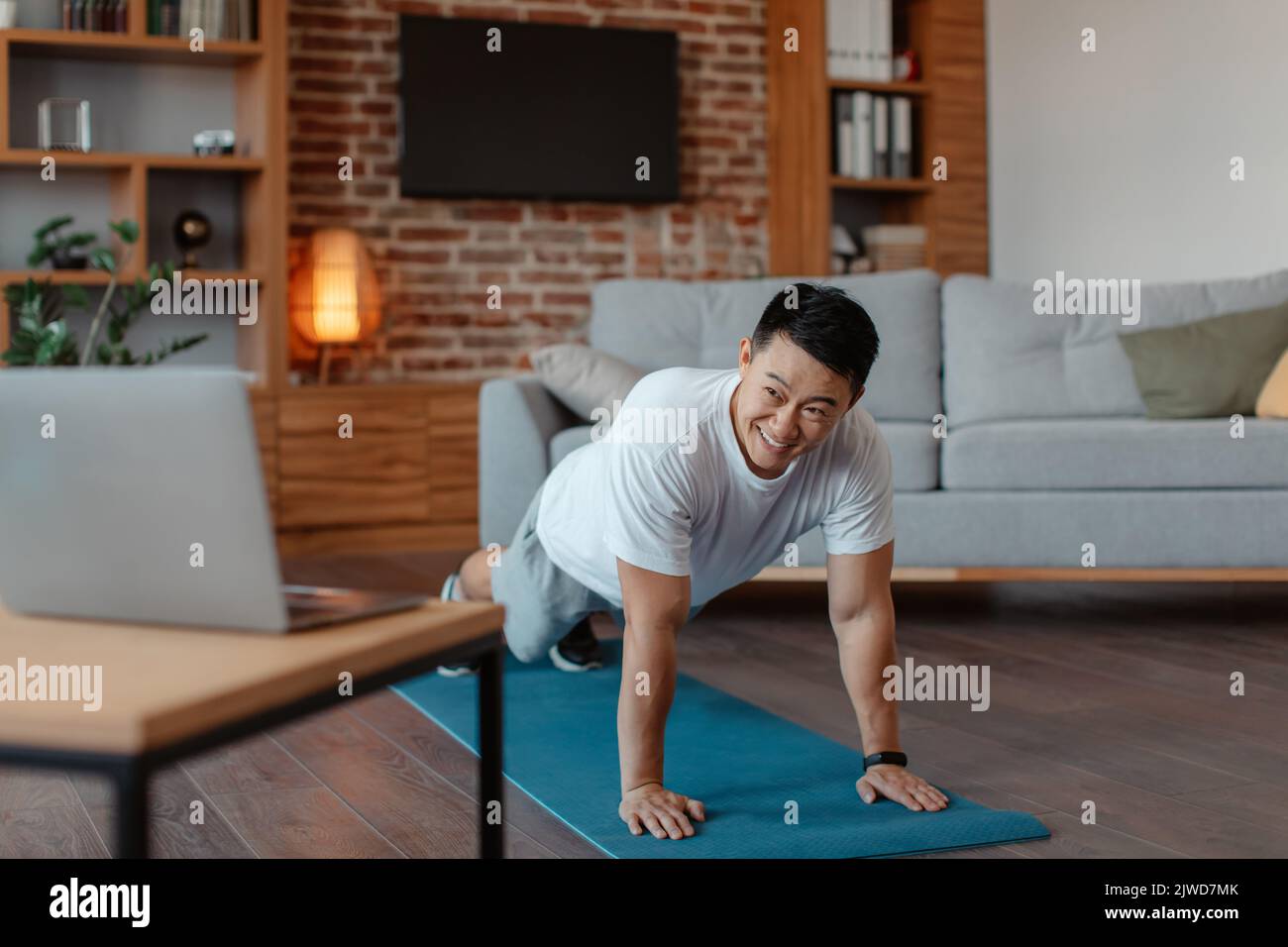 Concetto di classe di fitness online. Atletico uomo asiatico che plana sul tappetino yoga a casa, guardando video fitness sul computer portatile Foto Stock