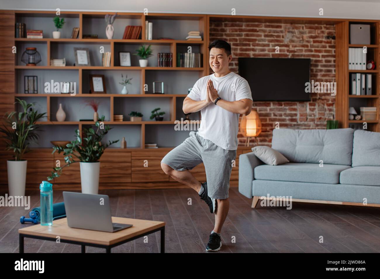 Uomo maturo coreano positivo che partecipa a una lezione di yoga online, in piedi in posizione albero di fronte al notebook, allenarsi a casa Foto Stock