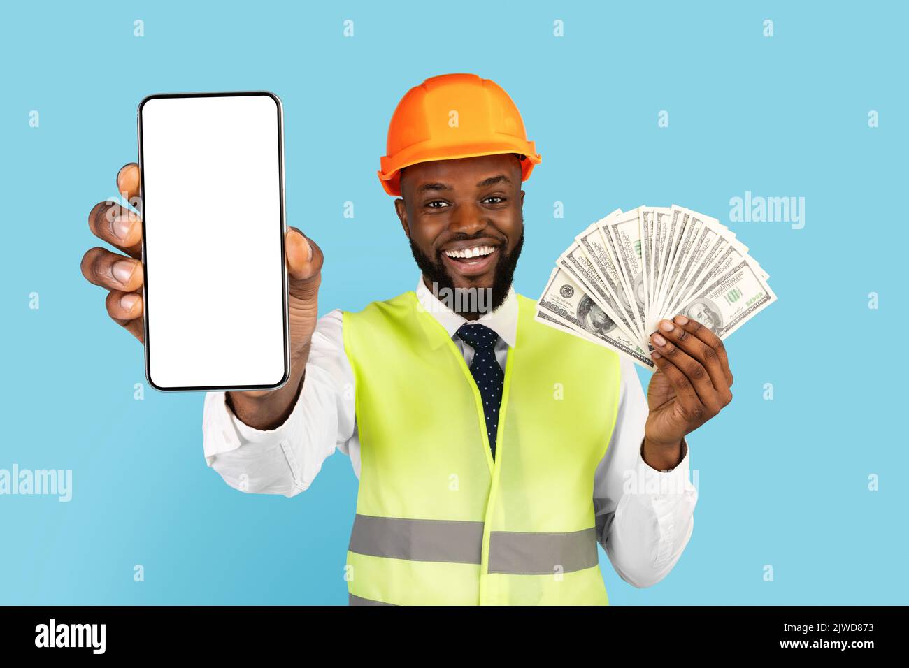 Uomo nero in berretto duro e giubbetto che tiene soldi e smartphone vuoto Foto Stock