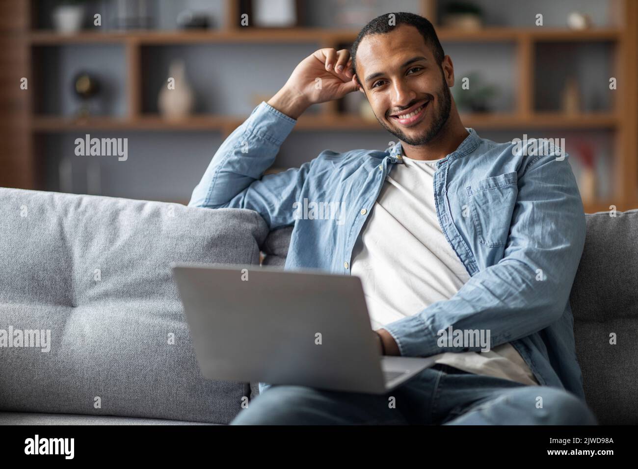 Bel maschio afro-americano che usa il portatile mentre si siede sul lettino a casa Foto Stock