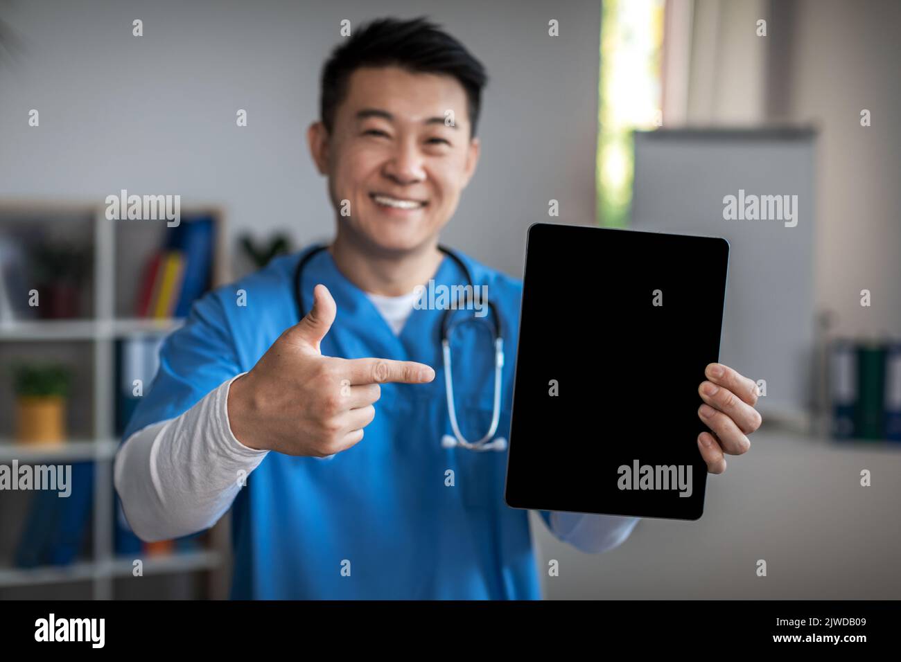 Allegro medico asiatico maturo uomo che punta il dito a tablet con schermo vuoto in clinica ufficio interno. Annuncio grande Foto Stock