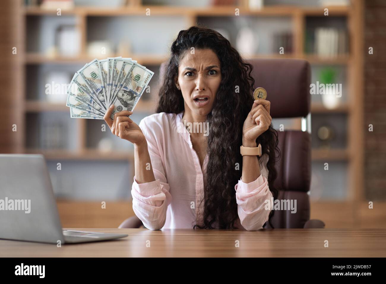 Arrabbiato donna d'affari in possesso di contanti e bitcoin, ufficio interno Foto Stock