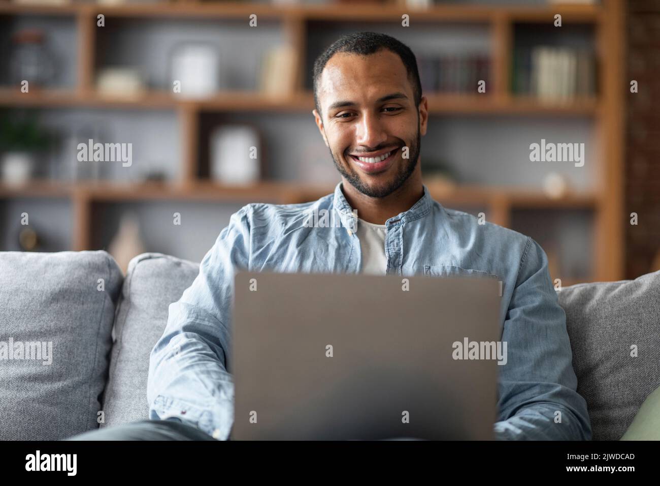 Comunicazione online. Bel giovane uomo nero seduto sul divano e utilizzando il computer portatile Foto Stock