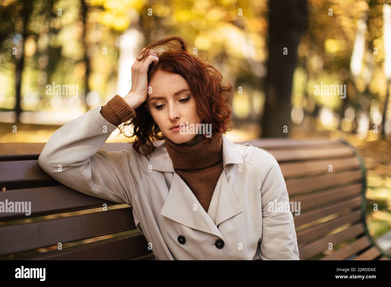 Triste stanco giovane signora europea in impermeabile siede su panchina, pensa, soffre di mal di testa e emicrania nel parco Foto Stock