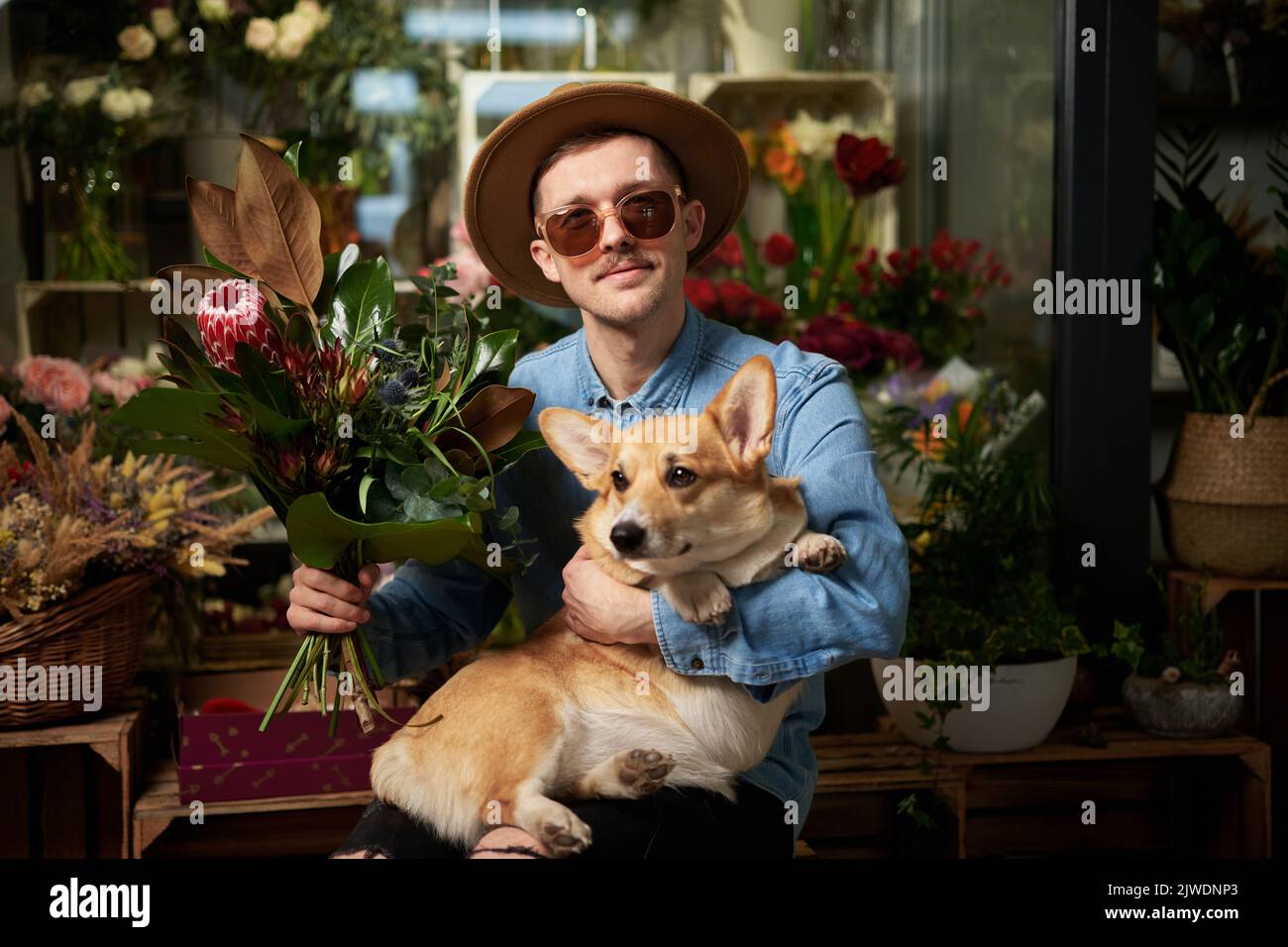Hippster uomo in occhiali da sole e cappello con bouquet di fiori freschi di primavera e corgi gallese cane in negozio di fiori. Giorno della mamma, giorno di San Valentino o Giornata internazionale della donna. Immagini di alta qualità Foto Stock