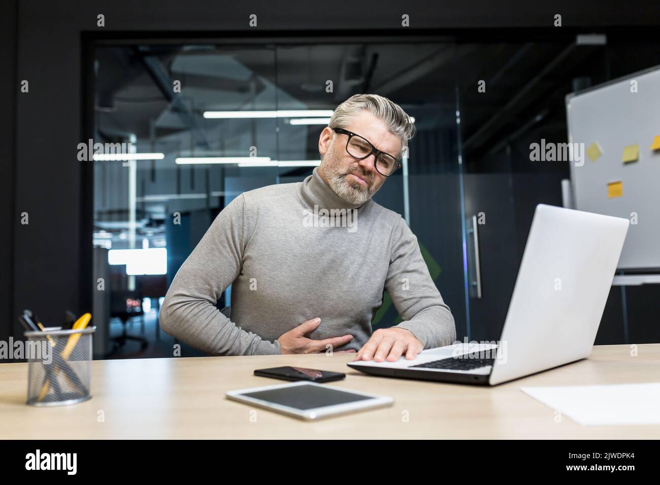 Malato uomo d'affari dai capelli grigi avvelenato, capo che tiene lo stomaco, avendo mal di stomaco grave uomo maturo che lavora in un ufficio moderno utilizzando un computer portatile. Foto Stock