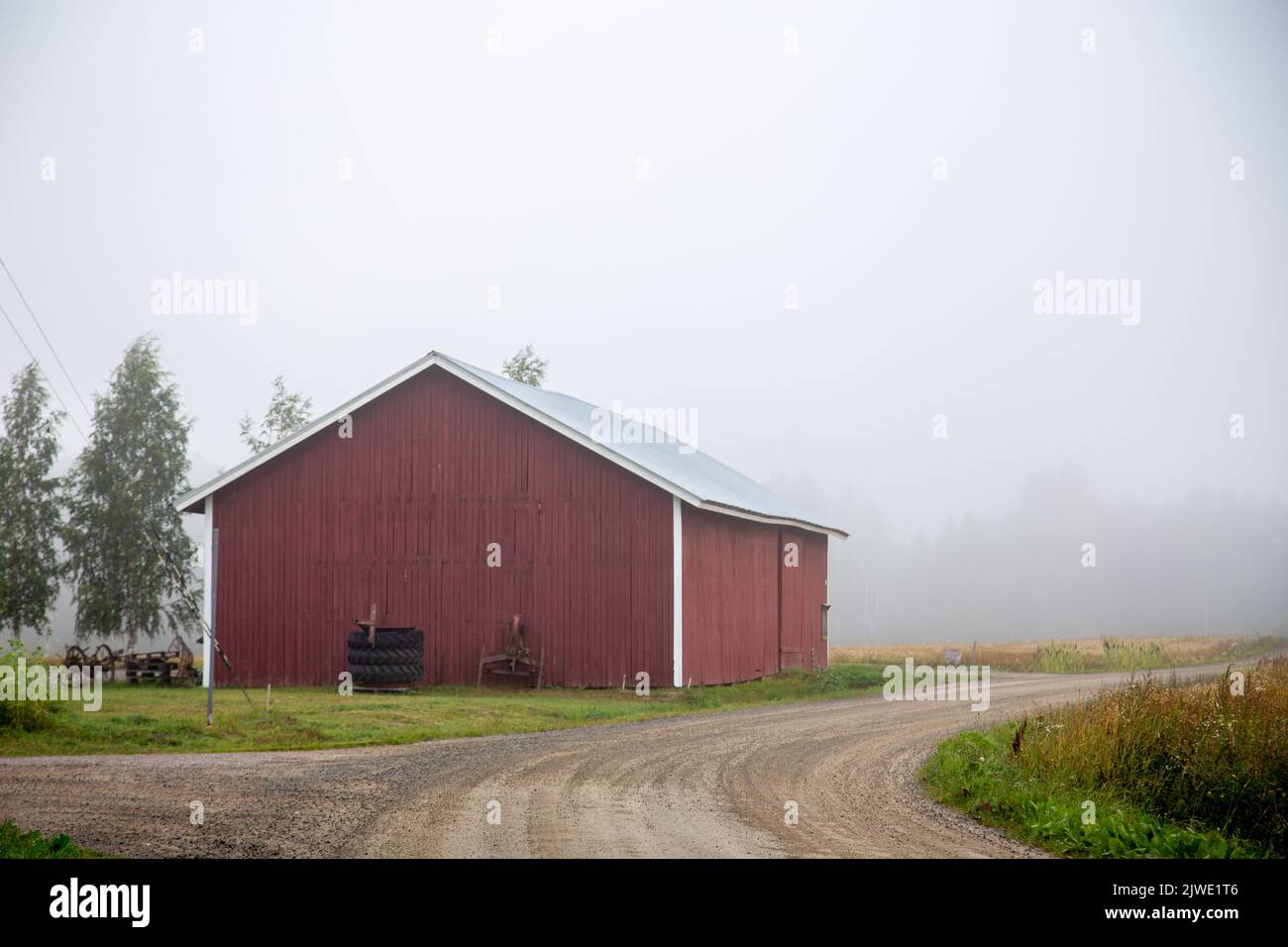 Un grande edificio agricolo rosso, una tranquilla strada del villaggio con sabbia, nebbia sullo sfondo. Foto Stock