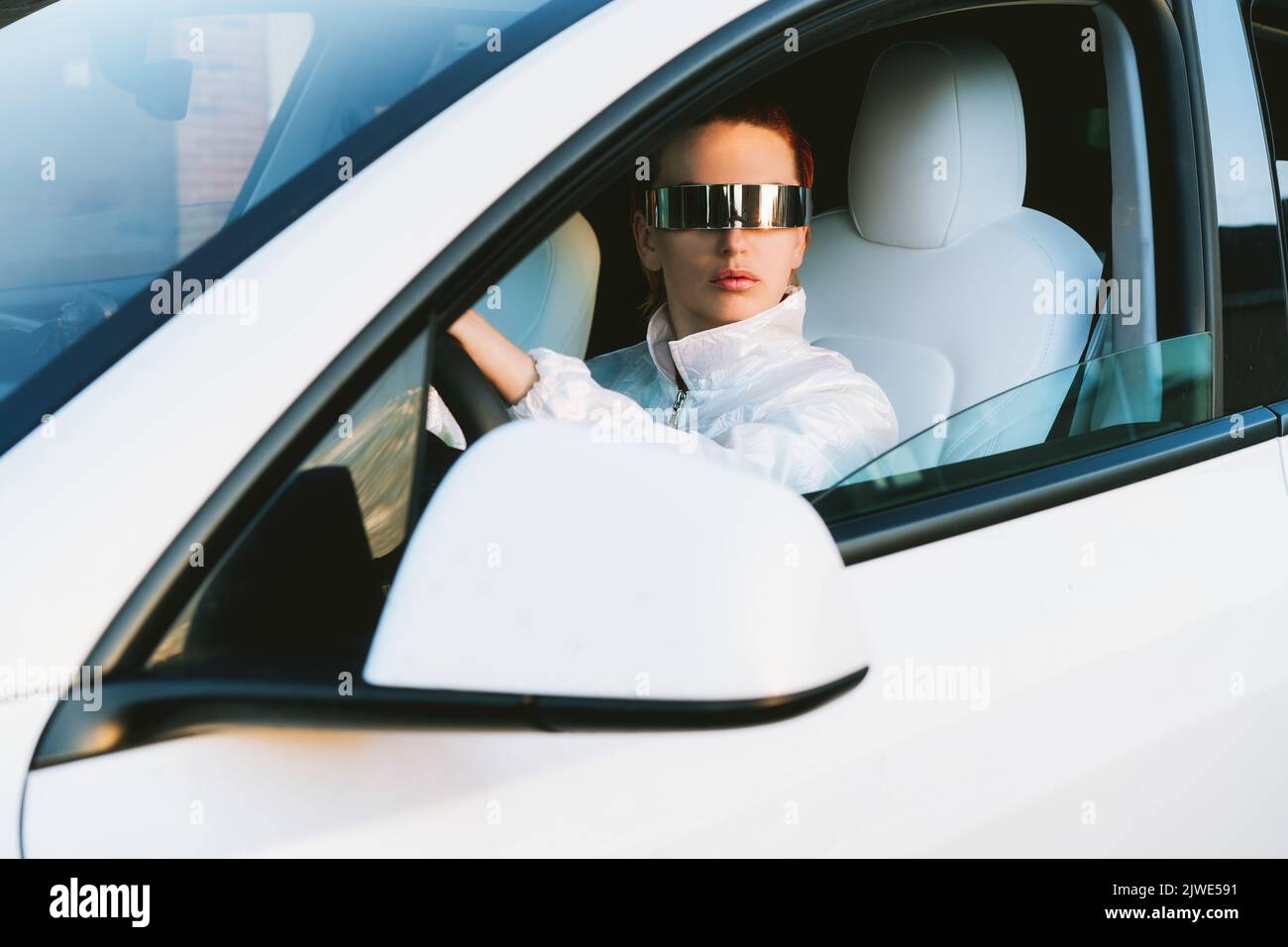 Ritratto di moda cyberpunk ragazza alla guida di una supercar bianco. Donna capelli rossi con occhiali futuristici si siede accanto al volante dell'auto. Futuro tecnolo Foto Stock