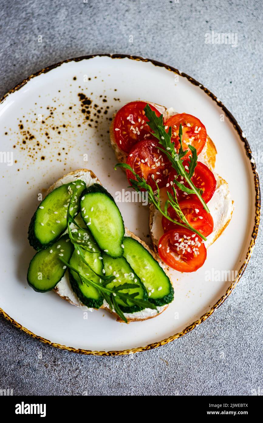 Vista dall'alto di due fette di pane tostato con formaggio cremoso, cetriolo, pomodoro, rucola e semi di sesamo Foto Stock