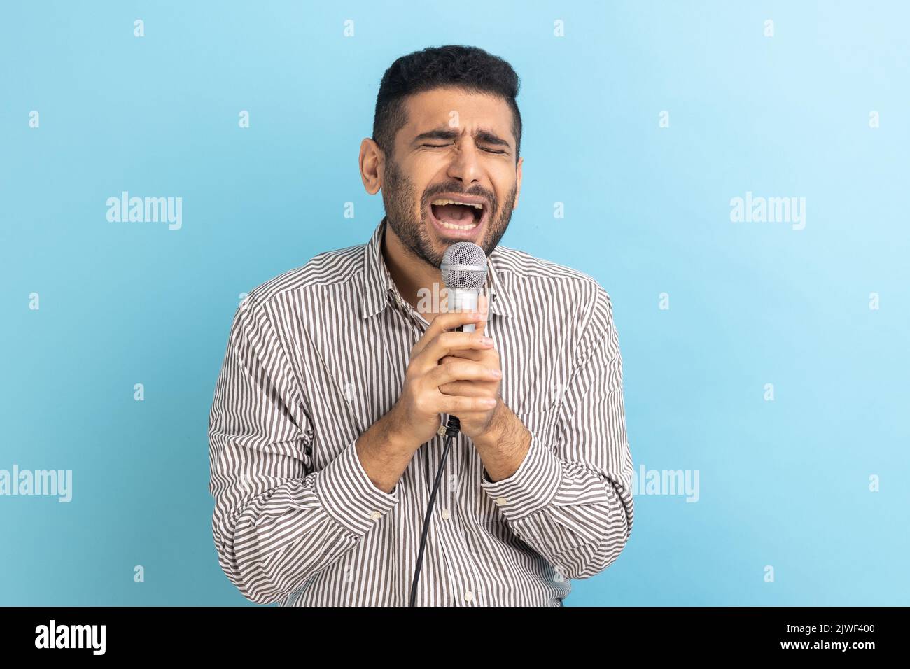 Ritratto di adulto bearded bel uomo d'affari cantando canzoni con microfono, prove prima dello spettacolo, indossare camicia a strisce. Studio in interni isolato su sfondo blu. Foto Stock