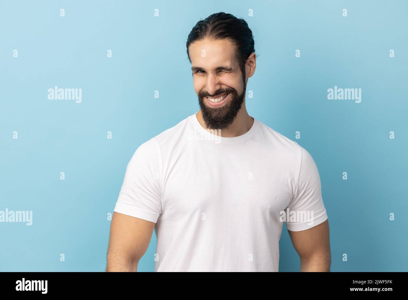 Ritratto di un uomo allegro con barba che indossa una T-shirt bianca di buon umore, sorridente in senso lato e che si agganita alla fotocamera con un sorriso toothy. Studio in interni isolato su sfondo blu. Foto Stock